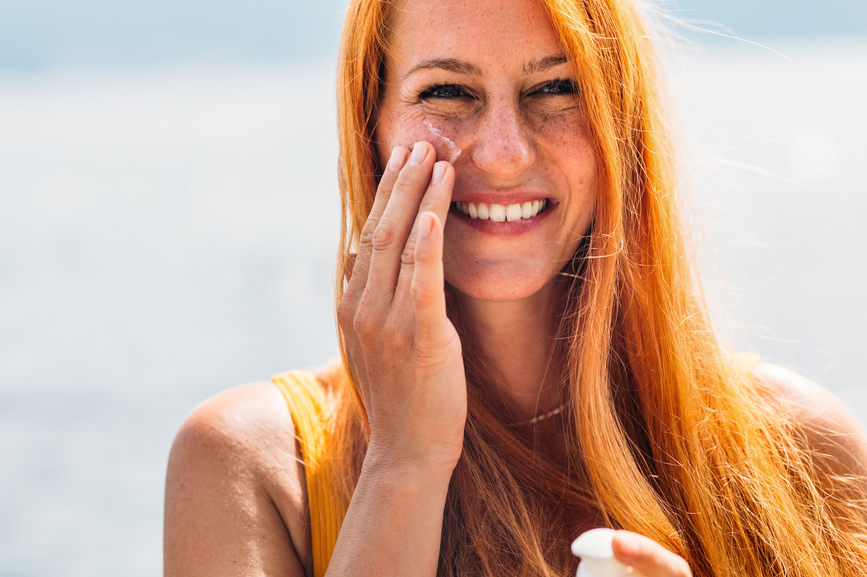Smiling redheaded woman applies sunscreen to her face while at the beach.