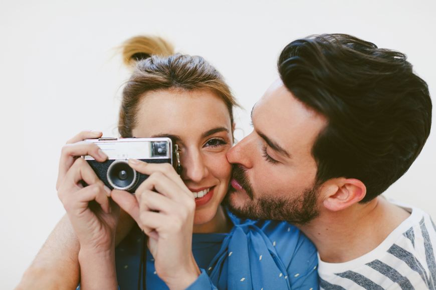 A man kisses a woman on the cheek as she holds up a camera.