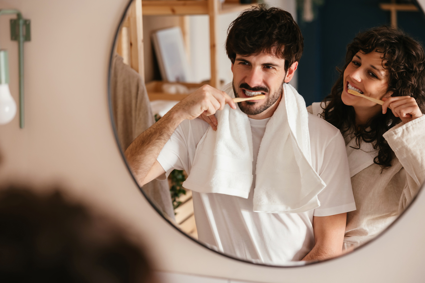 A boyfriend and girlfriend brushing their teeth while looking into the bathroom mirror.