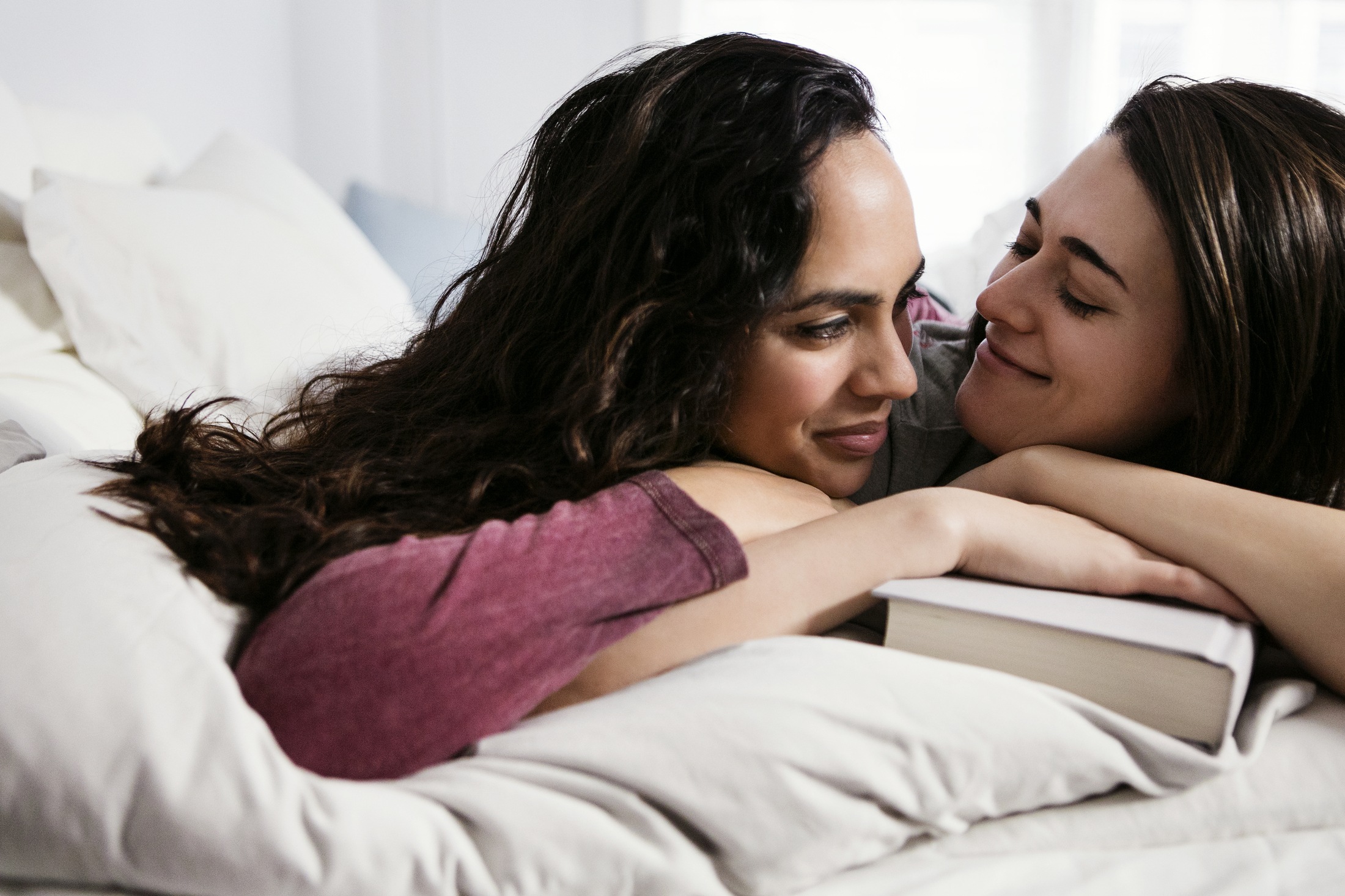 Two women lie on a bed and look into each other's eyes.