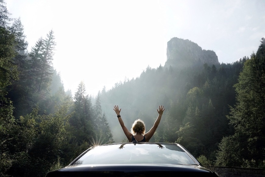 A woman raising her arms through the open sunroof of a car as it drives down a tree-lined path.