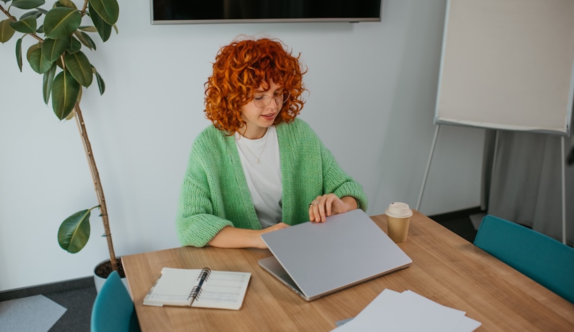 A young red-haired curly woman opens a laptop while sitting at her desk in the office