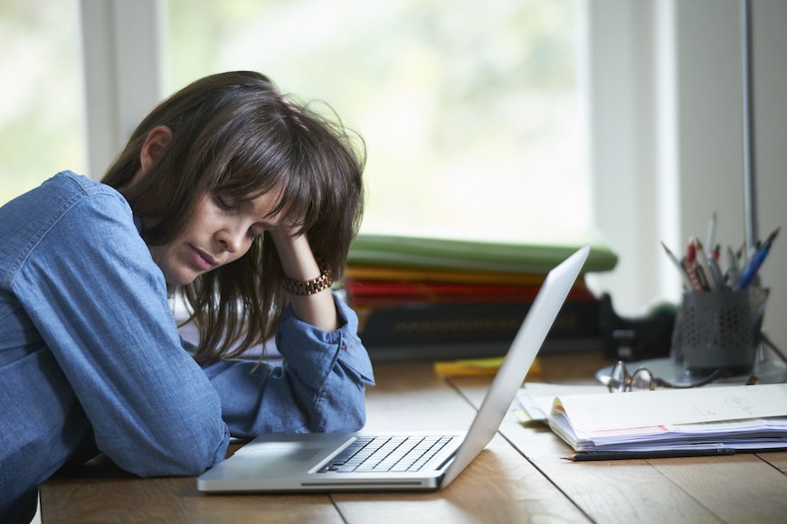 A woman holds her head in her hands and closes her eyes as she's on her laptop. She looks very tired.