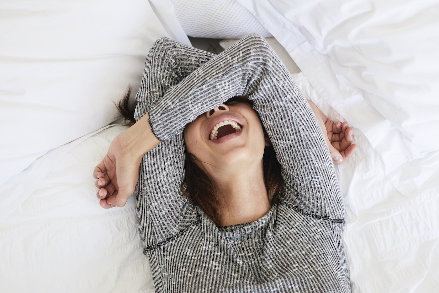 A woman wearing a gray shirt lies on a bed and smiles with her arms crossed over her head, covering her eyes.