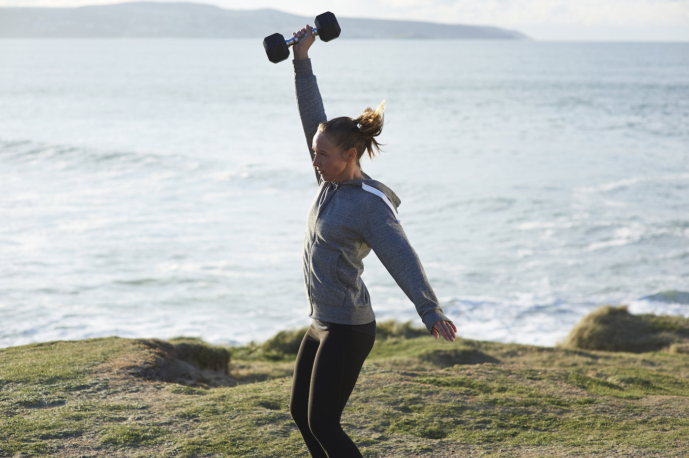 A woman standing on a cliff in front of the ocean raising a dumbbell above her head.