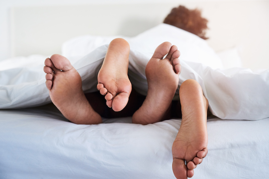 A view of the bottom of two peoples' feet as they lie in bed tangled in the sheets.