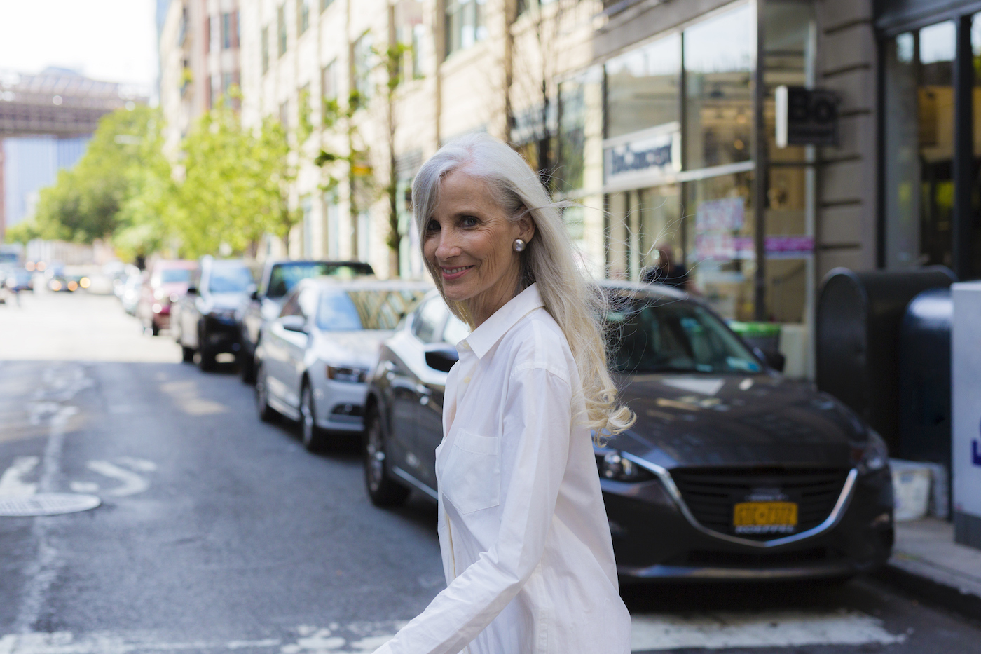 A woman with long gray hair and a white blouse turns back to the camera and smiles as she crosses a street in Brooklyn.