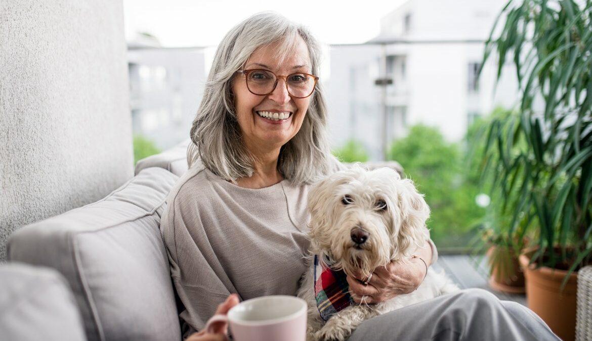 A smiling senior caucasian woman sits on a couch and holds her medium sized dog.
