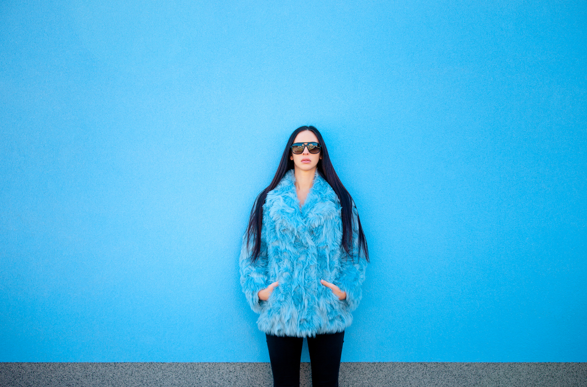 A woman in a blue furry coat and sunglasses stands against a blue background.