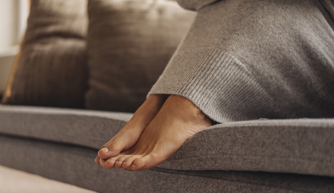 A woman's feet tucked into a long dress while sitting on the couch.