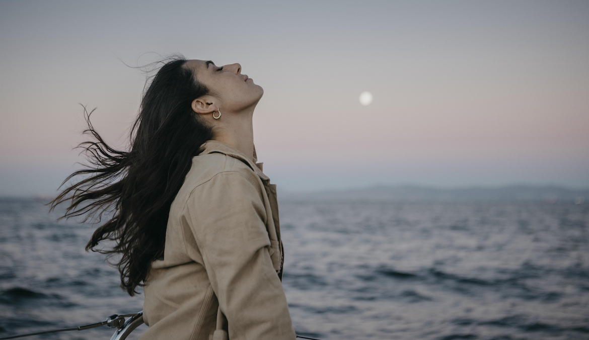 A woman throws her head back as she looks at the moon in the distance.