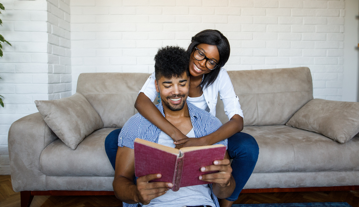 A couple sits and reads a book together. A female person sits on a couch behind the male person and wraps their arms around them.