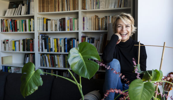 A smiling middle-aged woman sits on a chair in front of a large bookshelf and behind a healthy green plant, signifying the role of home design in longevity.