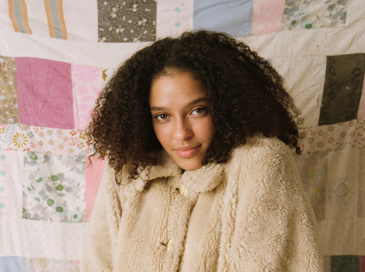 Portrait of a young woman with curly hair wearing a vintage coat posing in front of a quilt background, with her hands in her pockets, in pastel colors.