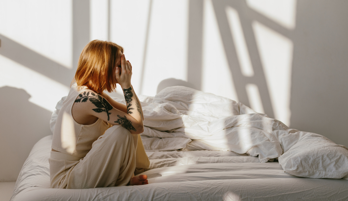 A redheaded woman sits on a bed with her head hidden in her hands, symbolizing the effects of not having sex.