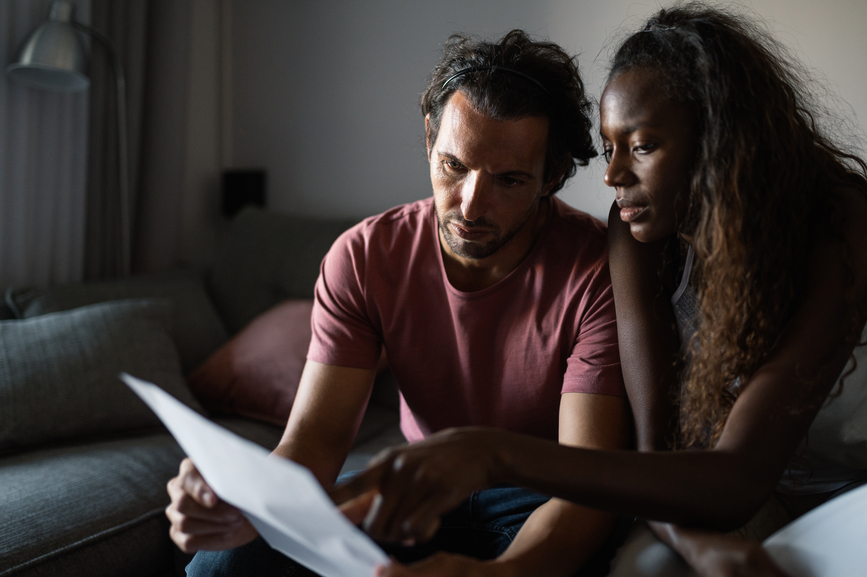 multiracial couple in casual wear sitting together on sofa and reading invoices with attention in living room in apartment