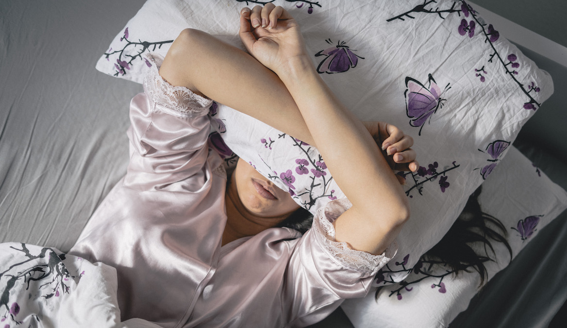 A woman lies in a bed with floral sheets and places a pillow over her face.