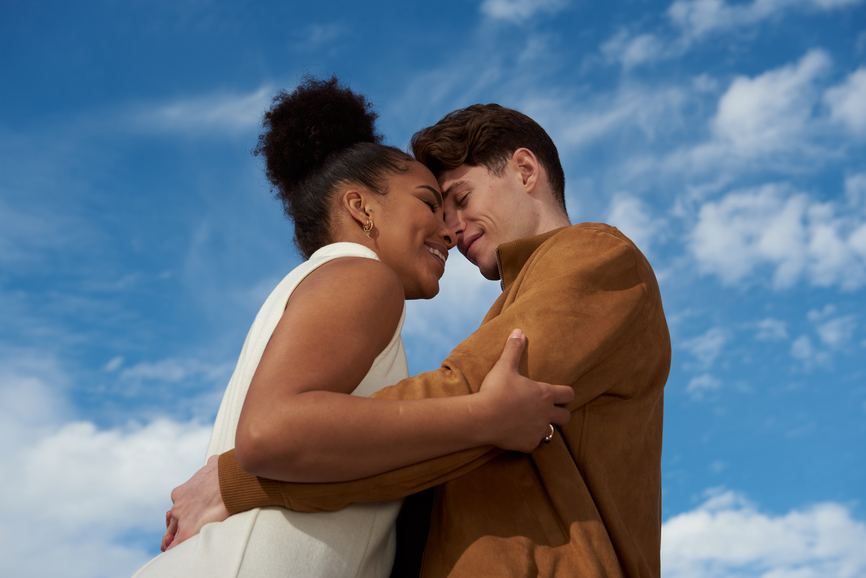 nterracial couple smiling, hugging with eyes closed outdoors with a blue sky