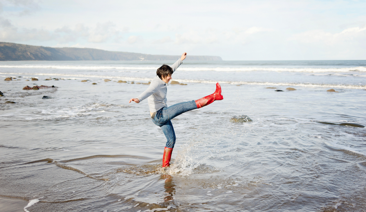 A woman red waterproof boots splashes around along a shoreline.