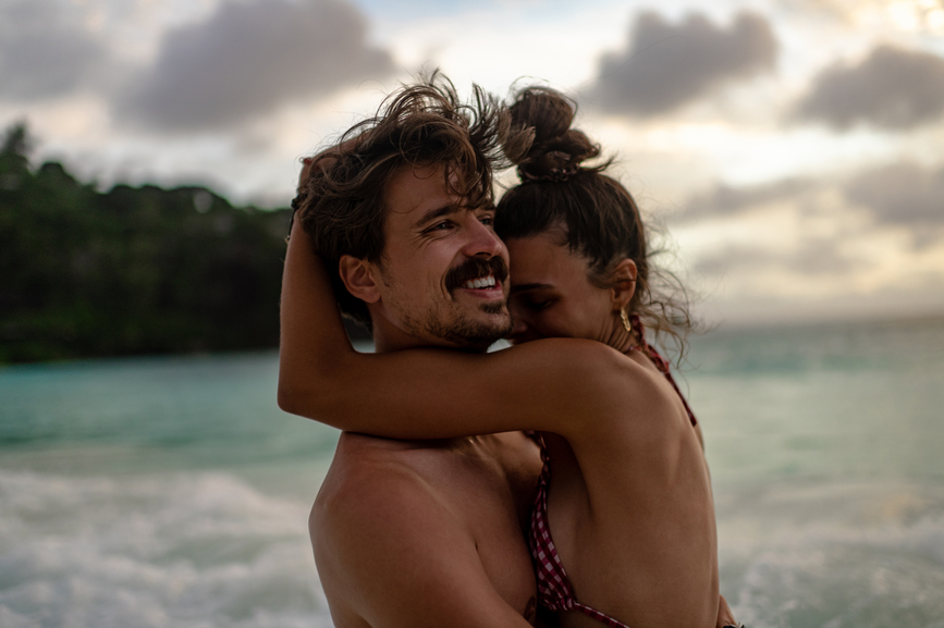 A man and woman embrace on the beach with a cloudy sky in the background.