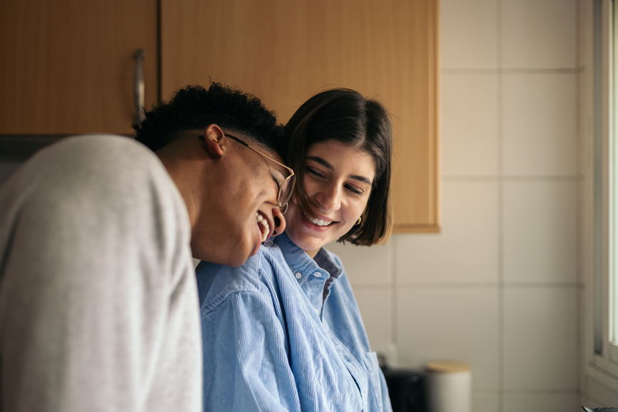 A woman leans on her partner in the kitchen as both smile.