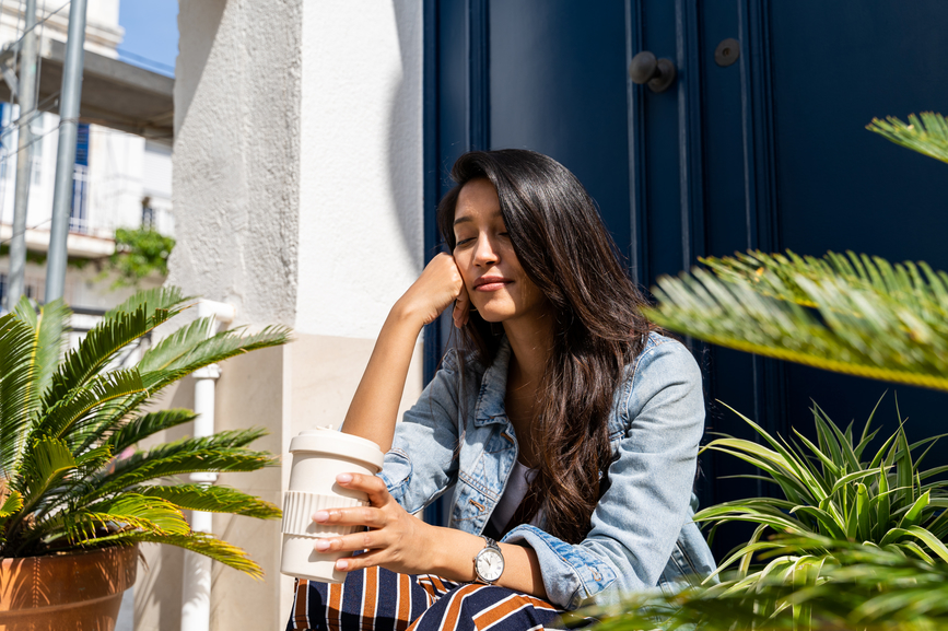 Pensive young woman thinking of future while sitting on doorway in front of door next to plants holding reusable coffe cup with closed eyes during a sunny day