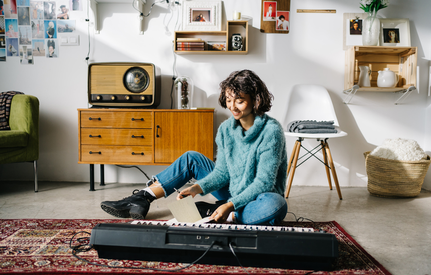 Calm thoughtful young woman in blue casual clothes standing beside electronic piano and thinking while writing new song in living room in retro style