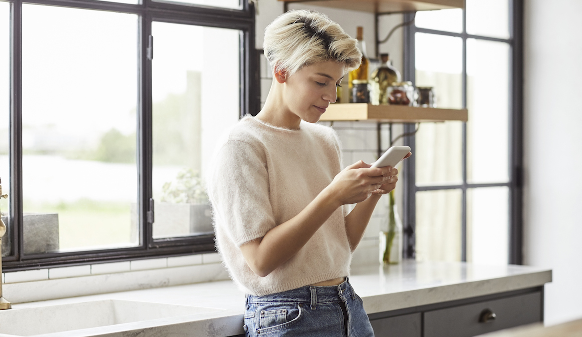 Smiling lesbian woman using smart phone. Female is text messaging while leaning on countertop. She is at home.