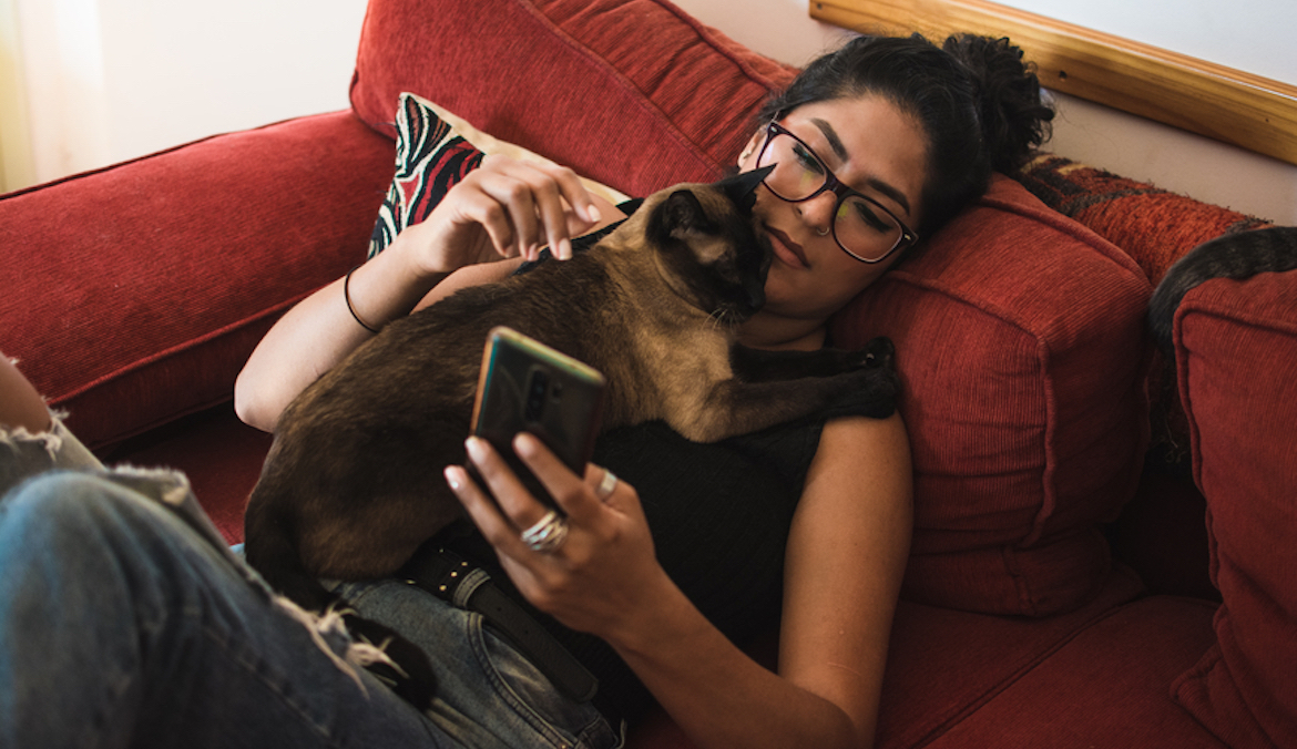 Woman sitting in her living room cuddling with her siamese cat while using her phone.