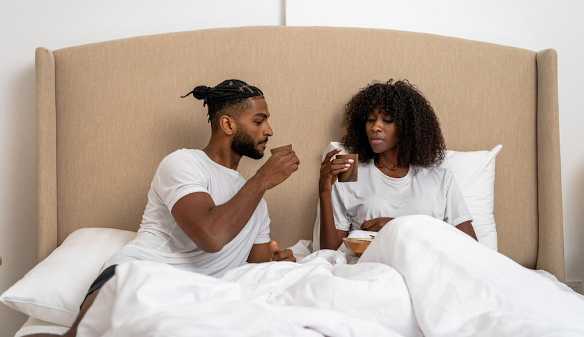 Young and attractive couple talking and having tea along with breakfast in the bed while sitting under white bed sheets.