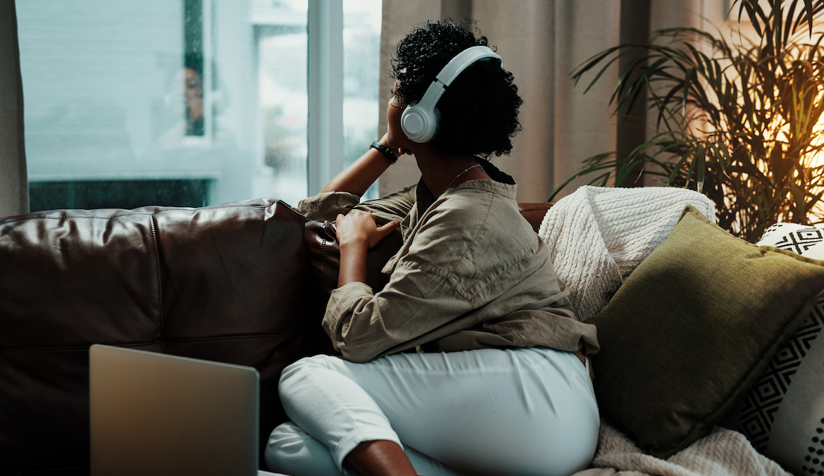 Shot of a young woman wearing headphones while looking out her living room window