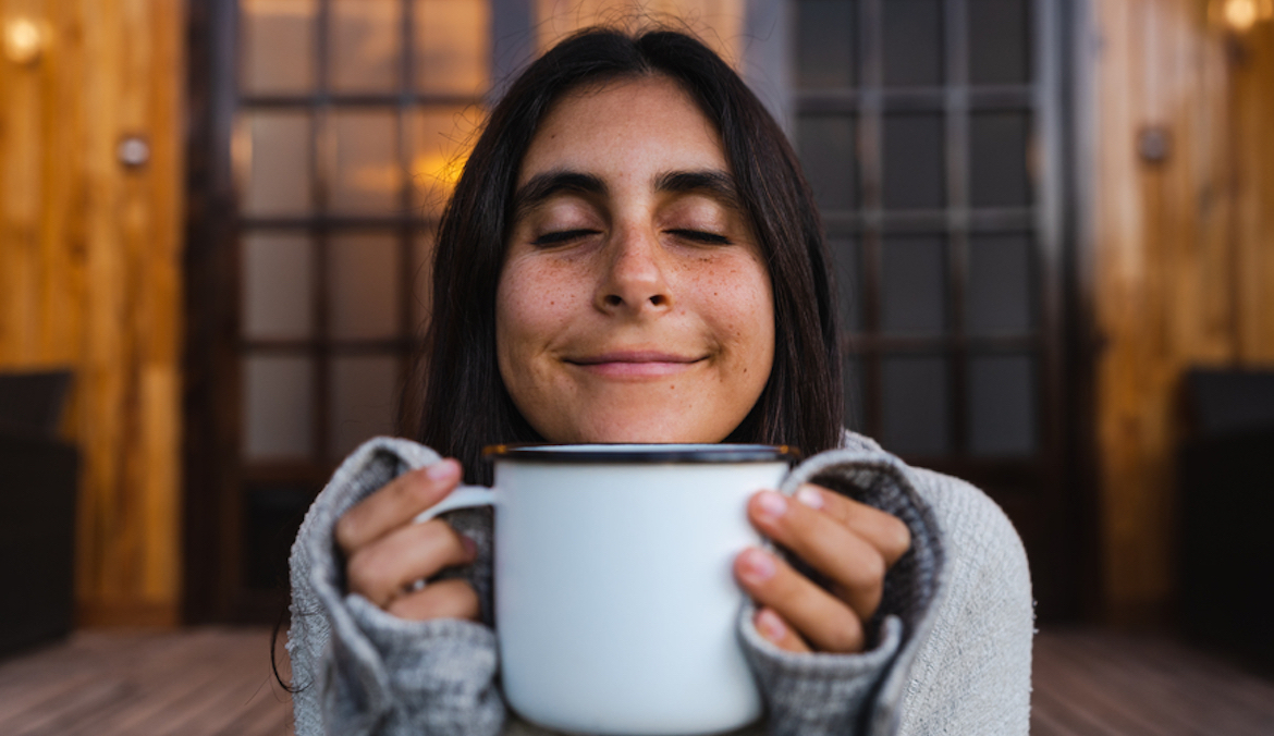 happy woman smiling with her eyes closed smelling her morning coffee outside a wood cabin