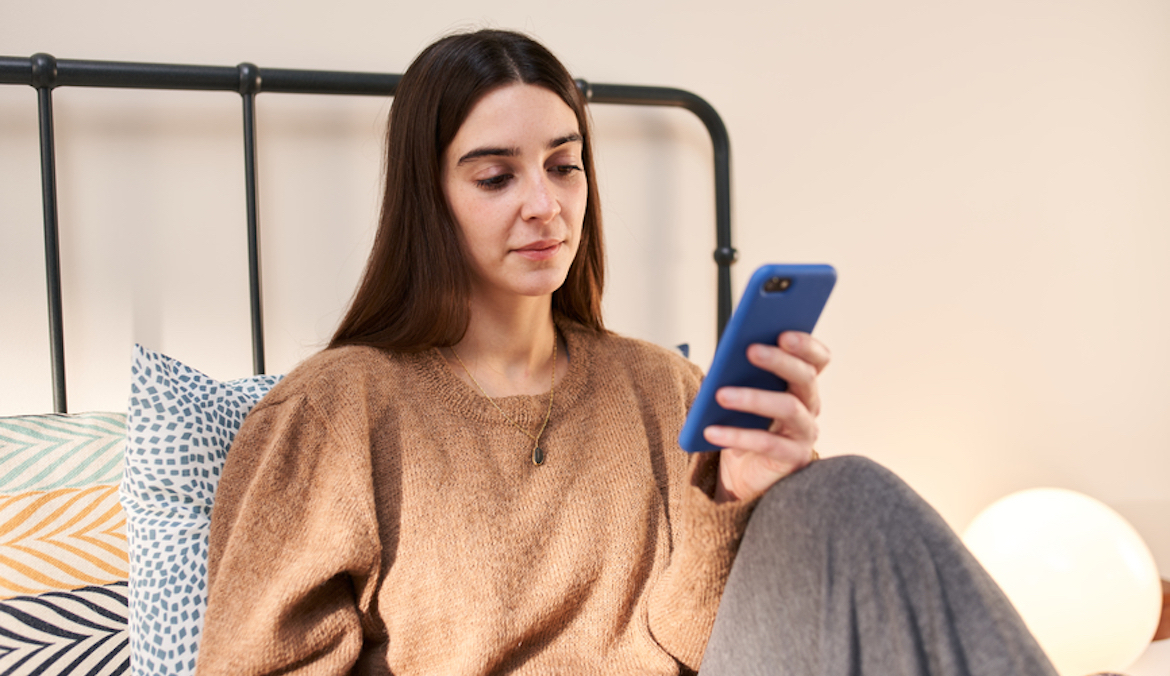 One young brunette female in warm brown sweater using cell phone while sitting in bed leaning on pillows.