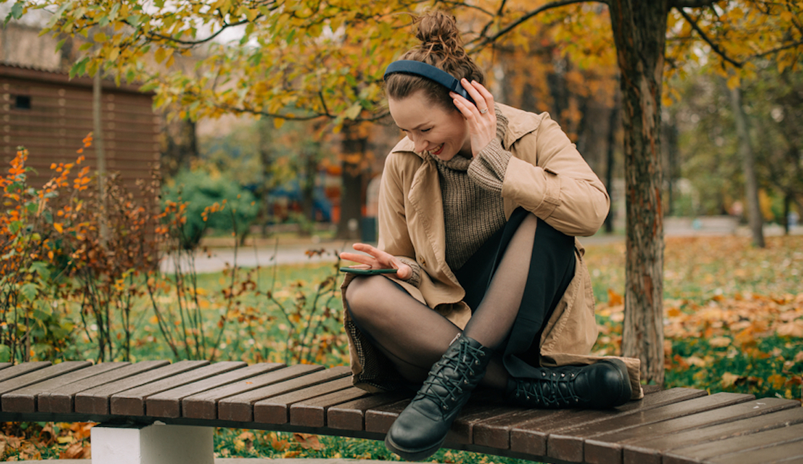 Happy woman using her phone while sitting on a bench and listening to music in autumn park
