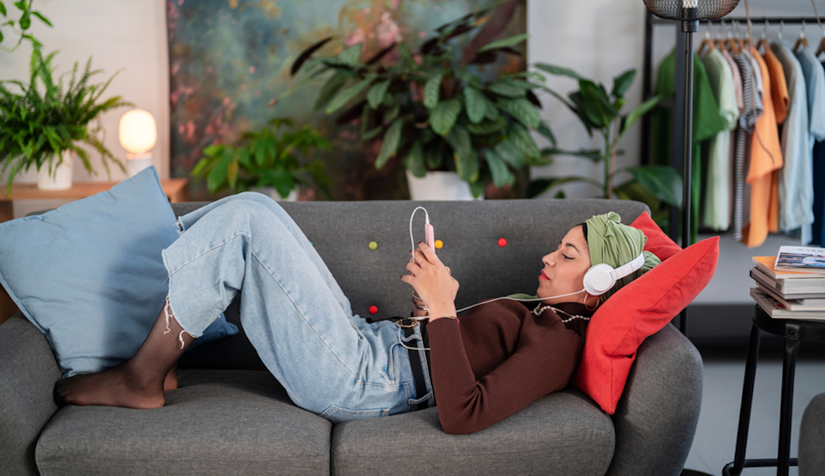 Side view of young Muslim female in casual clothes and h using mobile phone and listening to music while relaxing on sofa in living room.