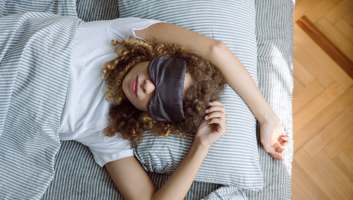Portrait of a curly young woman in a sleep mask in bed with concise bedding. Top view of sleeping young woman in bed with cotton bed sheets