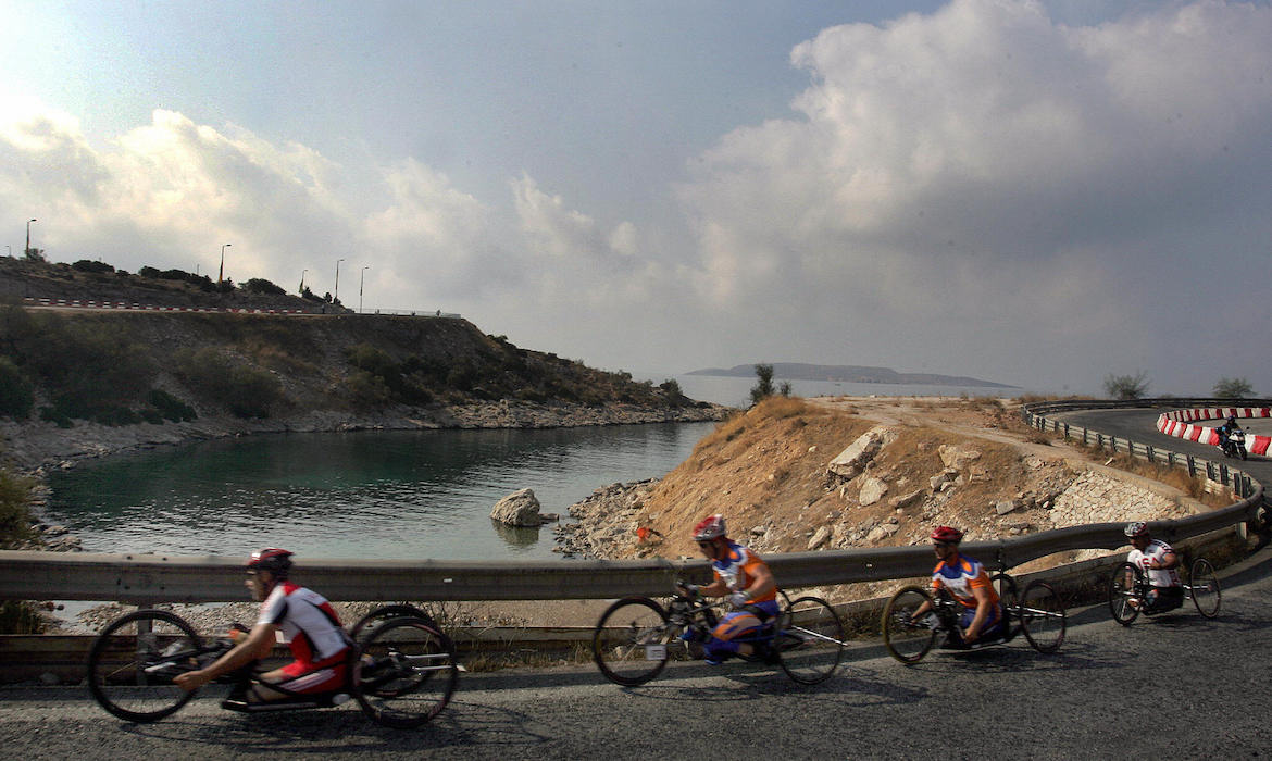 Handcyclists race on an asphalt road running along a rocky coastline.