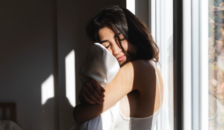 A pretty brunette woman hugs a pillow in a satin pillowcase.