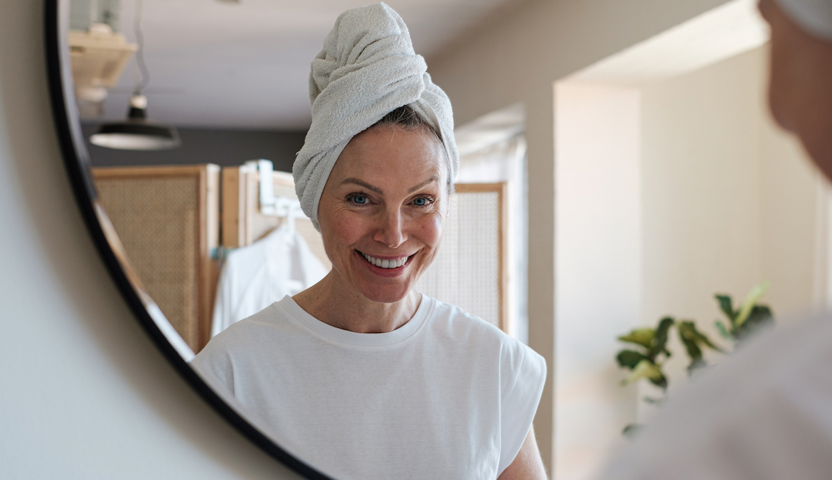 Portrait of a smiling blonde woman with a bath towel on her head looking at camera while is in skincare in front of a mirror in a bathroom