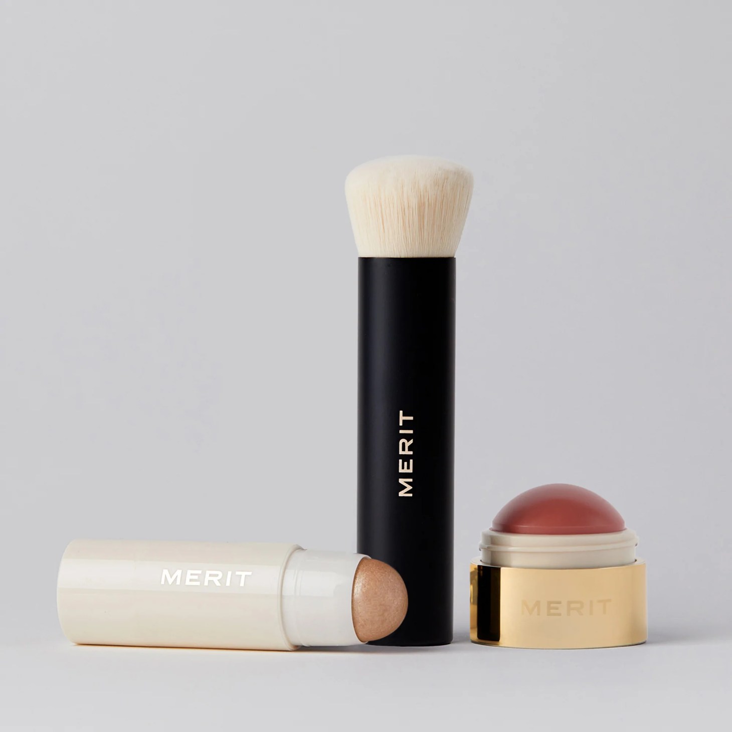 merit on the go set, which includes a blush balm, highlighting stick and blending brush, on a grey background