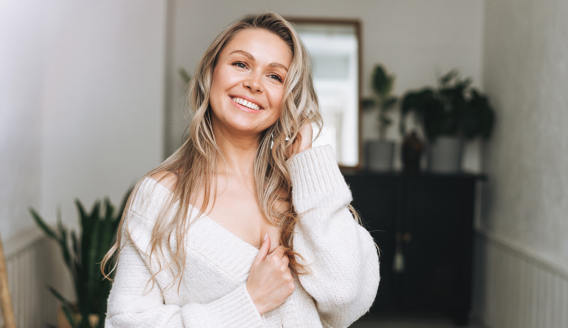 Blonde smiling woman 35 year clean fresh face and hands with long hair in cozy knitted cardigan at the bright interior