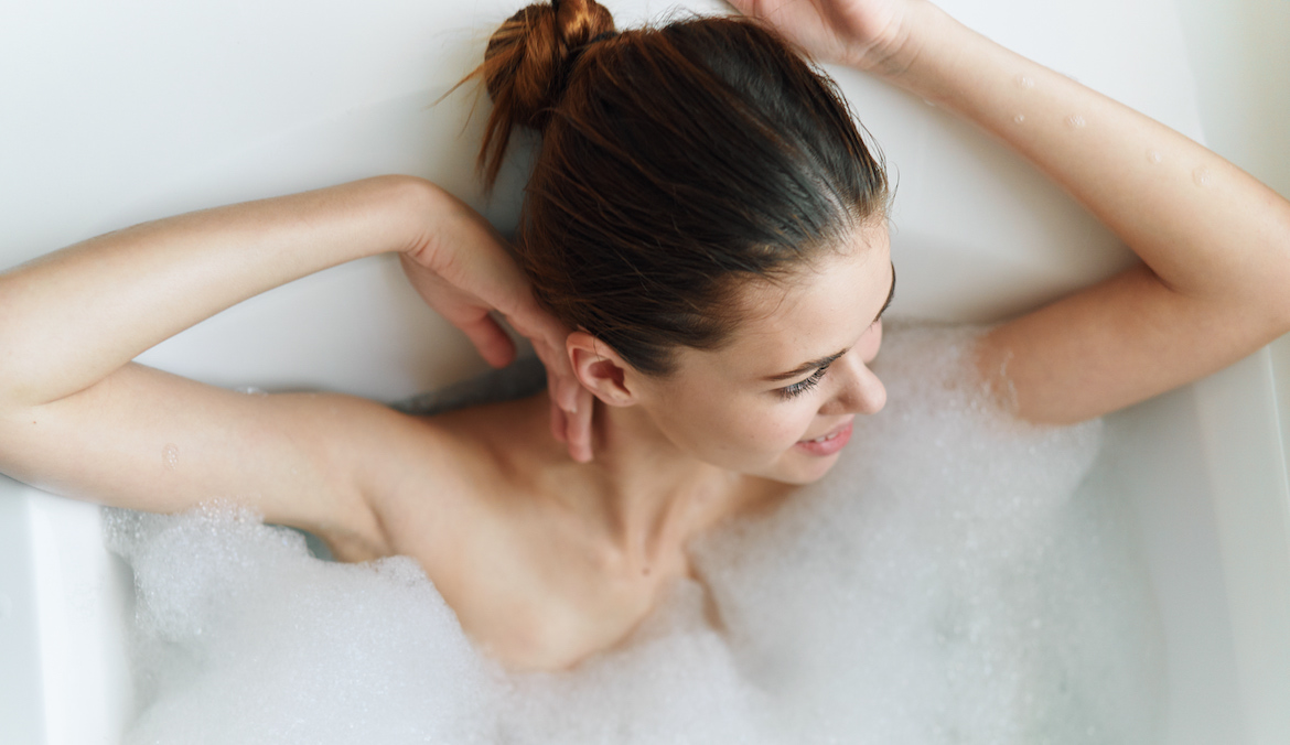 Smiling woman in bath tub with bubbles with her hair slicked back into a bun.