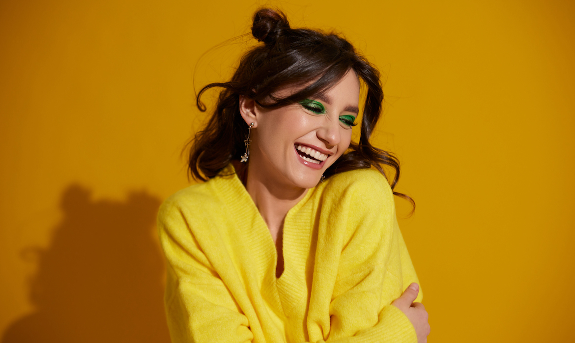 Brunette woman wearing space buns and green eye makeup smiling girl with bright makeup and wavy hair on a bright yellow background