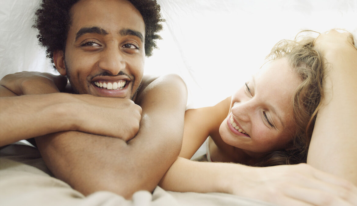 A young attractive couple lays on top of their bed underneath a bedsheet, smiling.