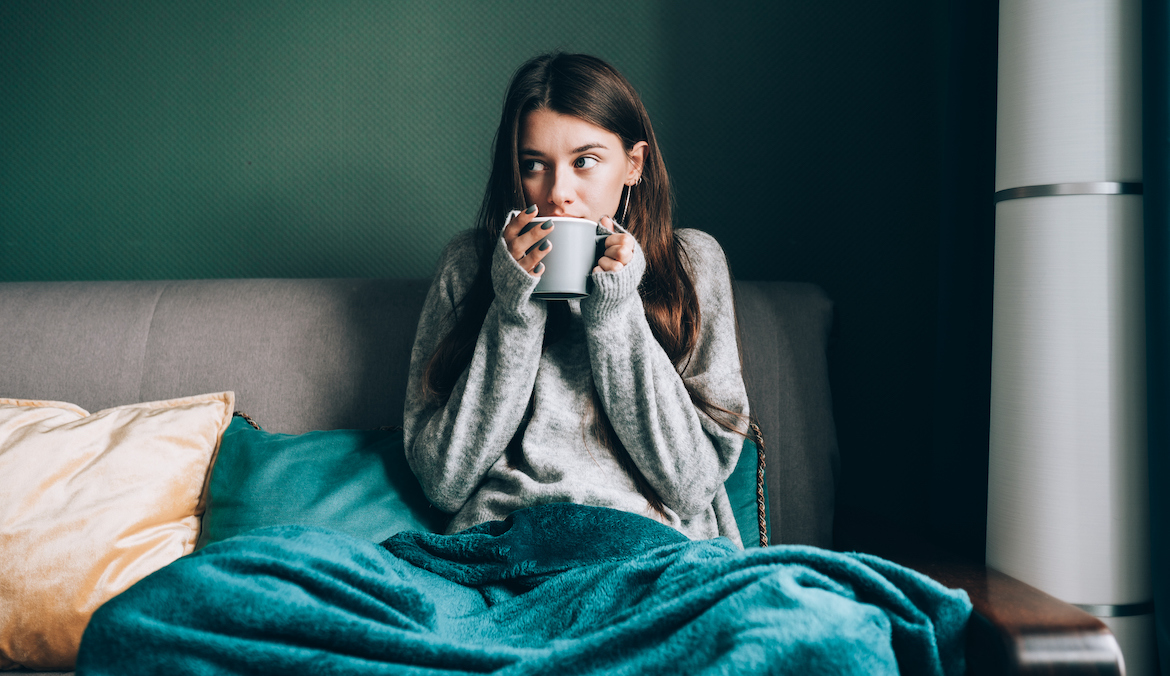 Shivering brunette woman in sweater, wrapped in duvet sitting on sofa and drink hot tea