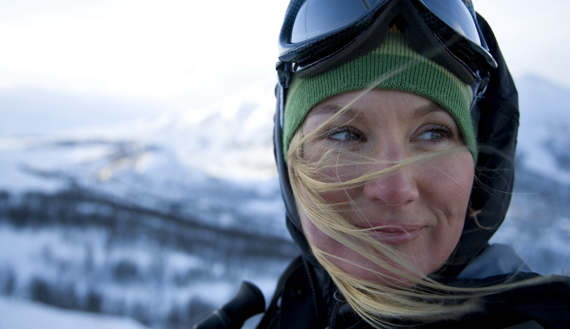 A woman with ski glasses looks to the side while hair blows across her face