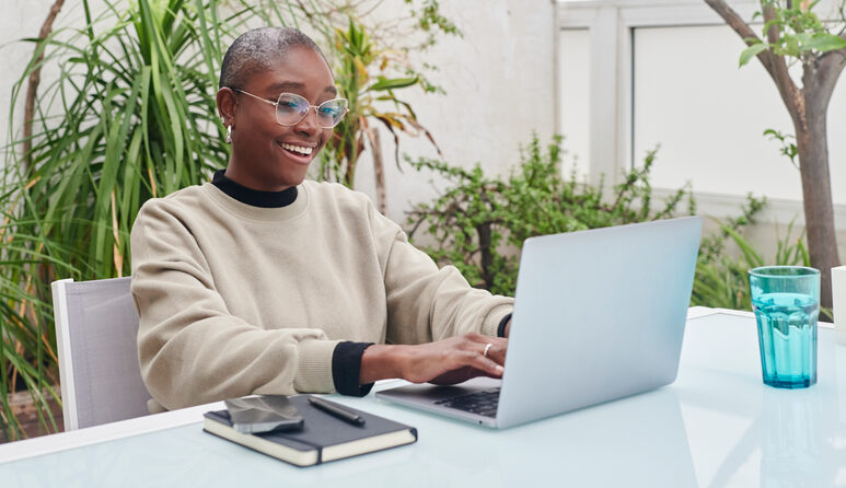 A young woman smiles while sitting outdoors typing on a laptop.