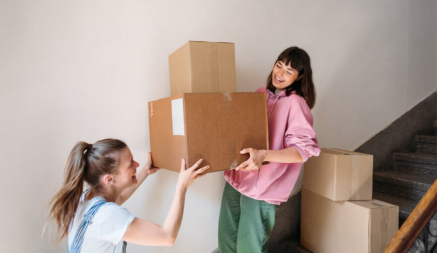 Two young women carry a pile of brown moving boxes together.