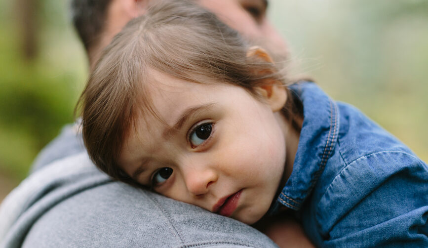 A young girl gives a forced hug to family member, resting her head on his shoulder while looking toward the camera.