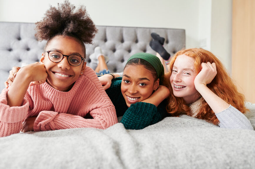 Group of three happy teens sitting on a bed smiling.